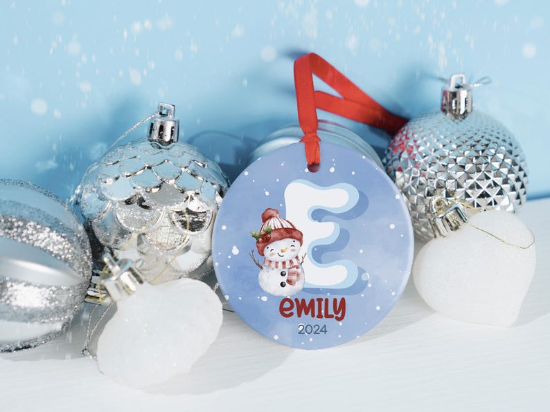 May include: A blue and white ceramic Christmas ornament with a snowman and the letter E. The ornament is personalized with the name "emily" and the year "2024".