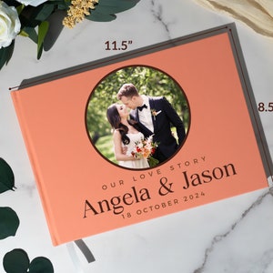 May include: A coral pink photo album with a circular photo of a couple in a park. The album is 11.5 inches wide and 8.5 inches tall. The text on the album reads "Our Love Story Angela & Jason 18 October 2024".