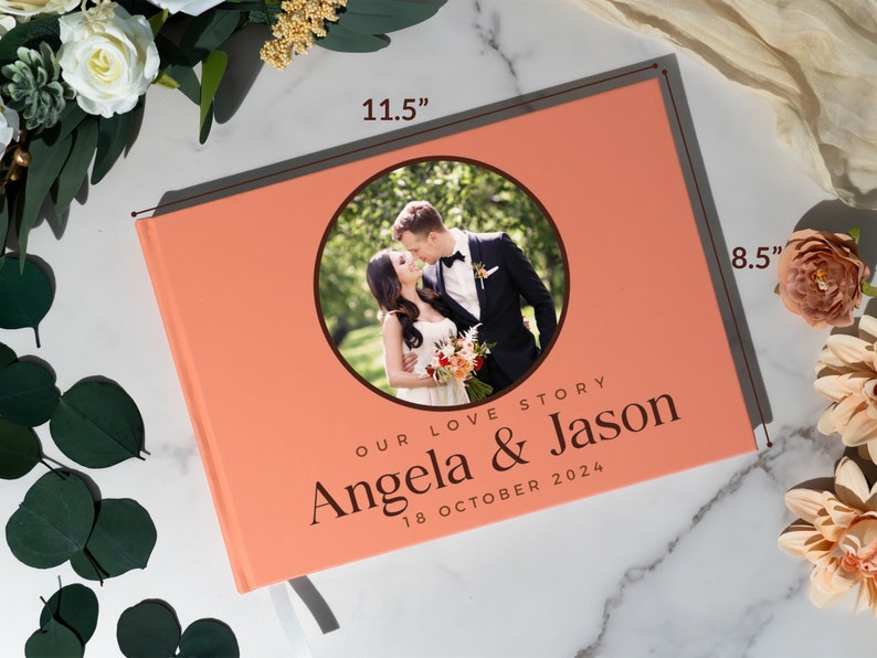 May include: A coral pink photo album with a circular photo of a couple in a park. The album is 11.5 inches wide and 8.5 inches tall. The text on the album reads "Our Love Story Angela & Jason 18 October 2024".