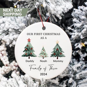 May include: White ceramic Christmas ornament with three watercolor Christmas trees, a red star, and the text "Our First Christmas as a Family of Three 2024".