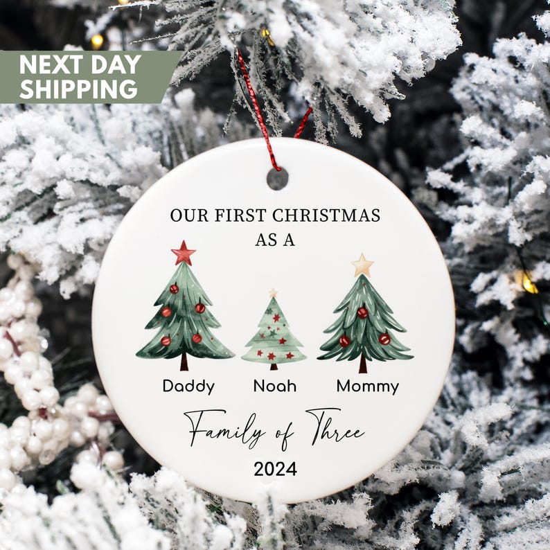 May include: White ceramic Christmas ornament with three watercolor Christmas trees, a red star, and the text "Our First Christmas as a Family of Three 2024".