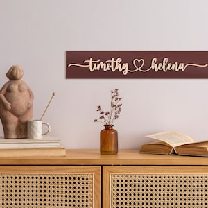 May include: A wooden sign with the names "timothy" and "helena" separated by a heart symbol. The sign is painted a deep burgundy color and is mounted on a white wall above a wooden dresser.
