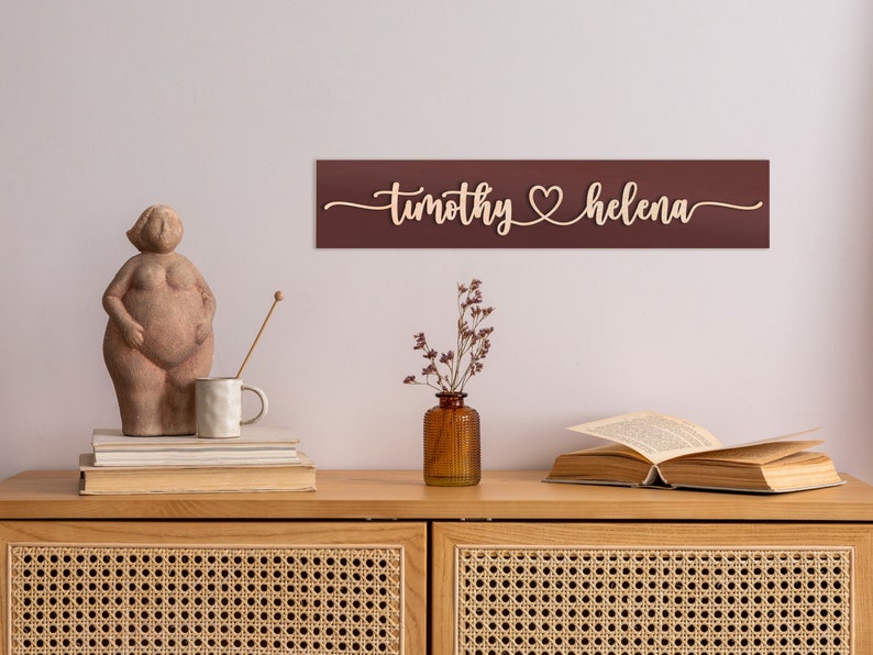 May include: A wooden sign with the names "timothy" and "helena" separated by a heart symbol. The sign is painted a deep burgundy color and is mounted on a white wall above a wooden dresser.