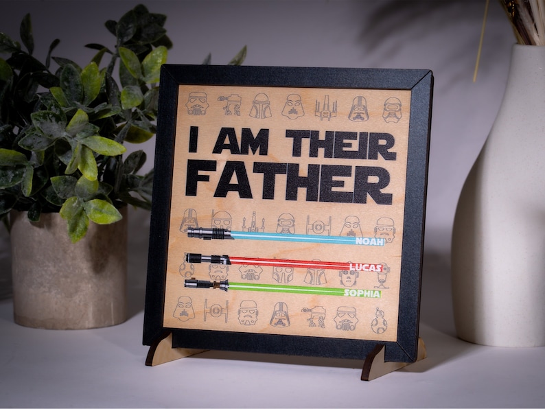May include: A framed wooden sign with the text "I AM THEIR FATHER" and lightsaber illustrations. The sign includes names below the lightsabers. The frame is black, and the background is a light brown color.