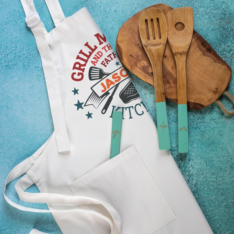May include: White kitchen apron with a red, white, and blue design. The apron has a pocket and the text "Grill Master and the Father of the Kitchen" with the name "Jason" in a banner. The apron is next to a wooden cutting board with two wooden spoons with turquoise handles.