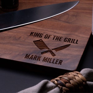 May include: A wooden cutting board with the text "King of the Grill" and "Mark Miller" engraved on it. Two crossed butcher knives are also engraved on the board.