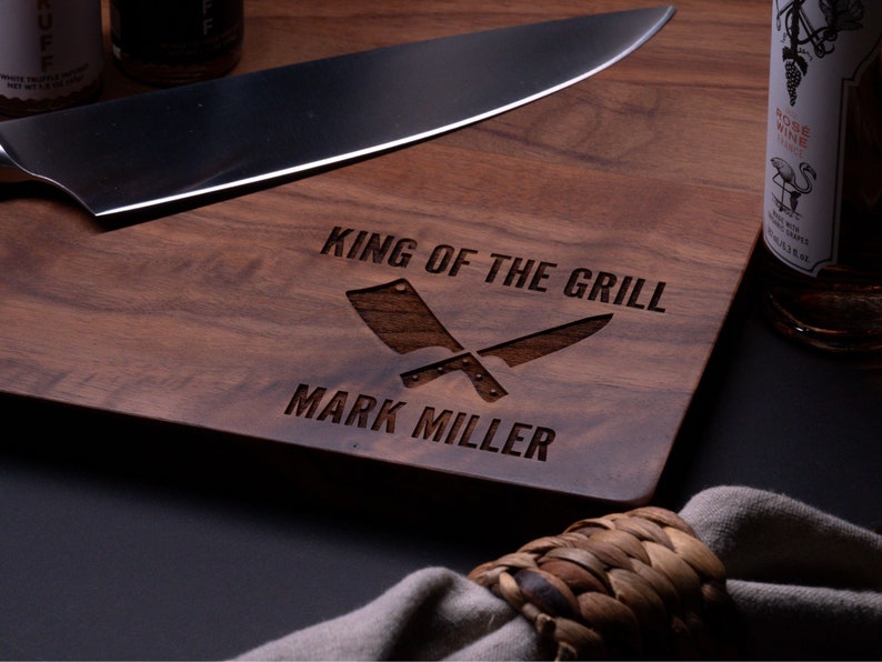May include: A wooden cutting board with the text "King of the Grill" and "Mark Miller" engraved on it. Two crossed butcher knives are also engraved on the board.
