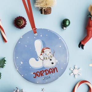 May include: A blue and white Christmas ornament with a snowman and the letter J. The ornament also has the name "JORDAN" and the year "2024" printed on it.