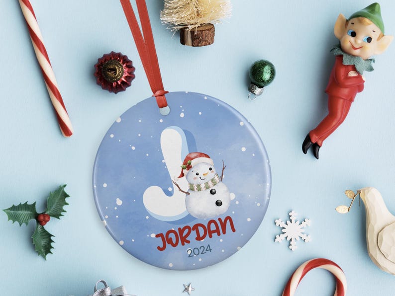 May include: A blue and white Christmas ornament with a snowman and the letter J. The ornament also has the name "JORDAN" and the year "2024" printed on it.