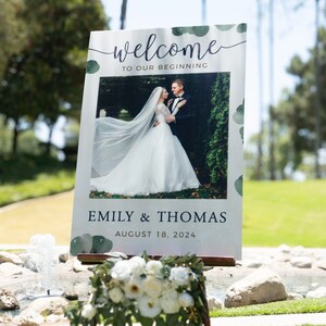 May include: A wedding welcome sign with the words "welcome to our beginning" in script. The sign features a photo of a couple and the names "Emily & Thomas" with the date "August 18, 2024". The sign is decorated with green watercolor accents.