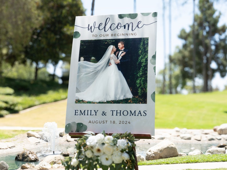 May include: A wedding welcome sign with the words "welcome to our beginning" in script. The sign features a photo of a couple and the names "Emily & Thomas" with the date "August 18, 2024". The sign is decorated with green watercolor accents.