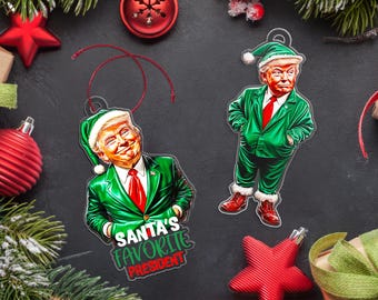 Op de afbeelding: Twee kerstversieringen met een karikatuur van een persoon in een groen pak en een kerstmanmuts. De versieringen zijn van metaal en hebben een rood lint. De tekst op een versiering luidt "Santa's favoriete president".