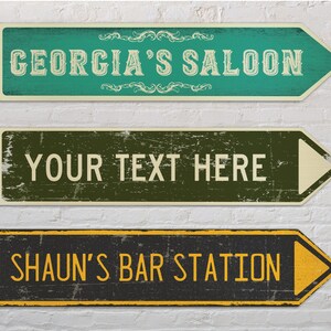 May include: Three vintage-style wooden arrow signs with distressed paint. The top sign is green with white text that reads "GEORGIA'S SALOON". The middle sign is green and brown with white text that reads "YOUR TEXT HERE". The bottom sign is black with yellow text that reads "SHAUN'S BAR STATION".