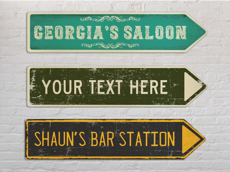 May include: Three vintage-style wooden arrow signs with distressed paint. The top sign is green with white text that reads "GEORGIA'S SALOON". The middle sign is green and brown with white text that reads "YOUR TEXT HERE". The bottom sign is black with yellow text that reads "SHAUN'S BAR STATION".