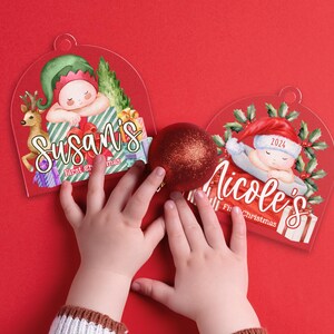 May include: Two arched acrylic ornaments featuring watercolor illustrations of babies in holiday attire. One shows a baby with an elf hat and reindeer, labeled Susan's First Christmas. The other shows a baby in a Santa hat, labeled Nicole's First Christmas.