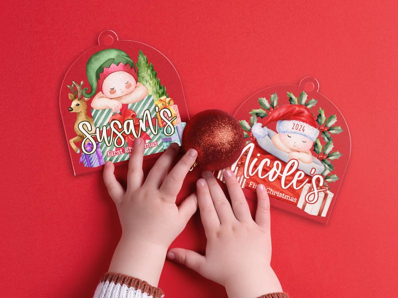 May include: Two arched acrylic ornaments featuring watercolor illustrations of babies in holiday attire. One shows a baby with an elf hat and reindeer, labeled Susan's First Christmas. The other shows a baby in a Santa hat, labeled Nicole's First Christmas.
