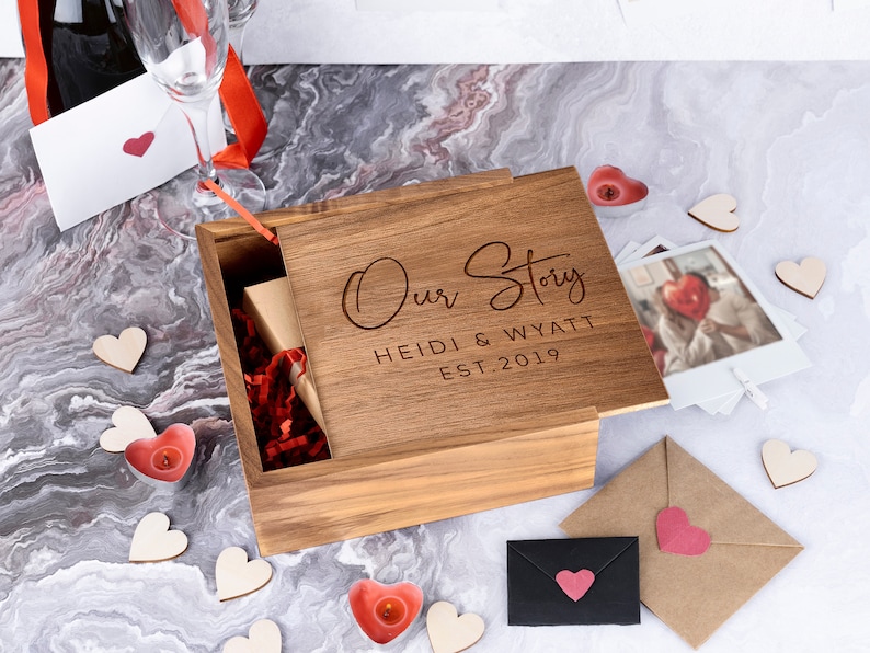 May include: A wooden box with a hinged lid, engraved with the text "Our Story Heidi & Wyatt Est. 2019". The box is open, revealing red tissue paper and a small brown box inside. The box is surrounded by small wooden hearts and red hearts.