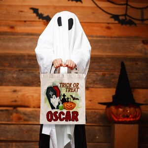 May include: A white ghost costume with a trick or treat bag. The bag is made of canvas and has a design of a graveyard, a ghost, a pumpkin, and the text "Trick or Treat Oscar".