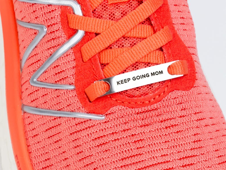 Puede incluir: Primer plano de una zapatilla de running rosa con un logotipo plateado y una etiqueta de metal plateado que dice "KEEP GOING MOM".