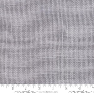 May include: A close-up of a light gray fabric with a woven texture. The fabric has a grid-like pattern, with a ruler along the bottom edge. The word "moda" is printed on the bottom edge.