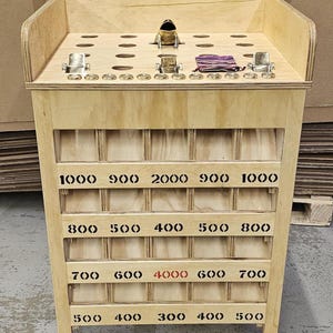 May include: A wooden tabletop game featuring a metal frog figure, multiple circular holes, and metal coin slots. The front panel displays four rows of numerical point values in black and red ink, ranging from 300 to 4000, organized into individual compartments.