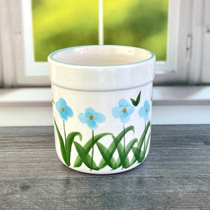 May include: White ceramic planter with a light blue rim and a hand-painted floral design. The design features light blue flowers with green stems and leaves. The planter is cylindrical and suitable for indoor or outdoor use.