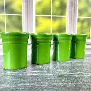 Könnte beinhalten: Vier leuchtend lindgrüne Keramikbecher, die in einer Reihe angeordnet sind. Jeder Becher hat einen glatten, zylindrischen Körper mit drei horizontalen Rillen, die den oberen Rand umschließen. Die Gefäße haben eine glänzende Oberfläche und eine einheitliche Farbe.