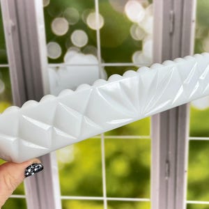 May include: A white, rectangular dish with a scalloped edge and a diamond pattern. The dish is made of opaque glass and is held in front of a window with a blurred green background.