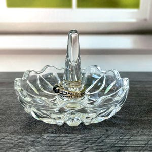 May include: A clear glass ring holder featuring a scalloped edge dish and a central conical post. A silver-toned ring with a dark stone and small clear accents is placed around the base of the central post of the glass ring holder.