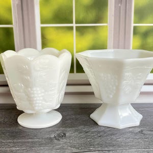May include: Two white milk glass vessels with embossed grape and leaf patterns. The left vessel features a scalloped rim and a circular pedestal base, while the right vessel has a hexagonal shape with a flared rim and a faceted pedestal base.