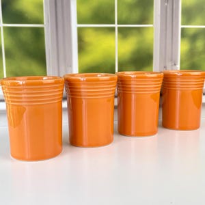 May include: Four orange ceramic tumblers with horizontal ridged detailing near the top. The tumblers are cylindrical and sit on a white surface, with a green background visible through a window.