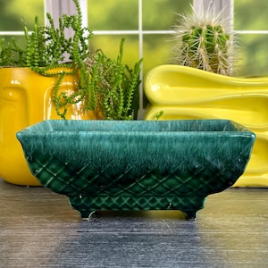 May include: A rectangular, dark green ceramic planter with a textured, diamond-patterned base and a smooth, glossy rim. The planter features four small feet and a deep, mottled glaze finish that transitions from a dark emerald to a lighter teal.
