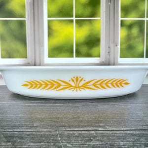 May include: A white, oval casserole dish with a yellow wheat pattern. The dish has a simple design with a slightly curved rim and is likely made of glass. The background features a window with a green, leafy view.