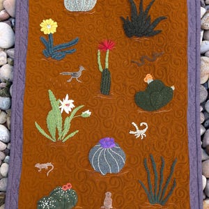 May include: A brown felt wall hanging with a desert theme. It features various cacti, a scorpion, a bird, and a lizard, all embroidered in different colors. The background is a brown felt with a swirling pattern.