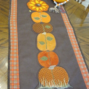 May include: A brown table runner with a plaid border features a stack of embroidered pumpkins with green leaves and a sunflower at the top. The runner is decorated with a brown, orange, and white color scheme.