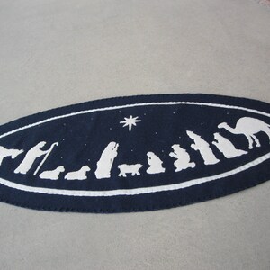May include: A navy blue oval-shaped felt table runner with a white embroidered scene of the nativity. The scene includes a star, a camel, a donkey, a shepherd, and the three wise men.