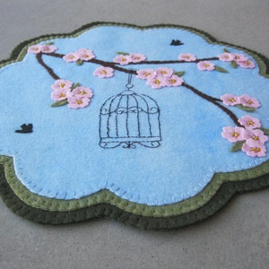 May include: A blue felt circle with a green border, featuring an embroidered design of a birdcage hanging from a branch with pink flowers. The flowers are embroidered with pink felt and have gold beads for centres.