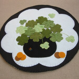 May include: A white felt circle with a black felt border and a scalloped edge. The center of the circle features a black felt pot with green felt shamrocks. Two orange felt circles are on either side of the pot.
