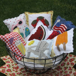 Könnte beinhalten: Ein Drahtkorb gefüllt mit bunten Stoffkissen mit sommerlichen Motiven, darunter ein Wassermelonenstück, ein Strandstuhl, ein Eis am Stiel, ein Getränk mit einem Schirm und eine amerikanische Flagge.