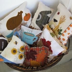 May include: A wicker basket filled with handmade felt pillows featuring Thanksgiving themes. The pillows have various designs including a turkey, a cup of coffee, a football, sunflowers, and fall leaves. The pillows are all white with colorful accents.