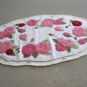 Könnte beinhalten: Weißer ovaler Teppich mit einem rosa-grünen Blumendesign. Der Teppich ist mit rosa Rosen und grünen Blättern bestickt.