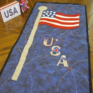 May include: A blue fabric wall hanging with a white star-spangled banner, the letters USA, and red, white, and blue fireworks.