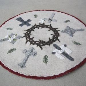 Könnte beinhalten: Ein weißer Filzkreis mit rotem Rand, der eine braune Dornenkrone, graue Kreuze und weiße Blumen zeigt. Das Design ist ein traditionelles christliches Symbol für die Passion Christi.