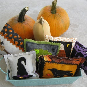 May include: A blue metal basket filled with handmade Halloween-themed fabric pillows. The pillows feature various designs, including a witch's hat, a broom, a raven, and a pumpkin. Two orange pumpkins are in the background.