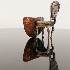 May include: A miniature silver chair with a brown, ornate back and a brown, wooden seat. The chair is sitting on a black surface.