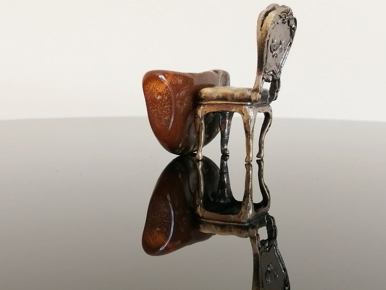 May include: A miniature silver chair with a brown, ornate back and a brown, wooden seat. The chair is sitting on a black surface.