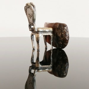 May include: A miniature silver chair with ornate details sits on a brown rock, creating a whimsical and surreal scene.