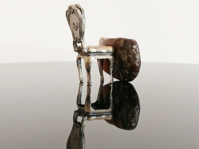 May include: A miniature silver chair with ornate details sits on a brown rock, creating a whimsical and surreal scene.
