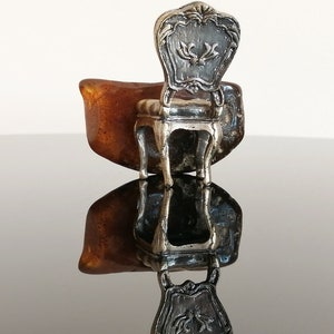 May include: A silver miniature chair with a detailed ornate back and a brown resin base. The chair is sitting on a reflective surface.