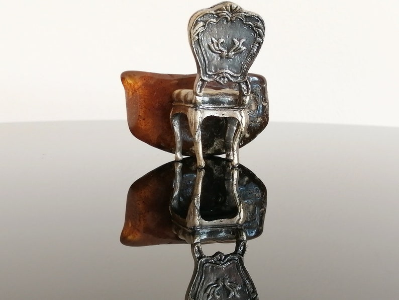 May include: A silver miniature chair with a detailed ornate back and a brown resin base. The chair is sitting on a reflective surface.